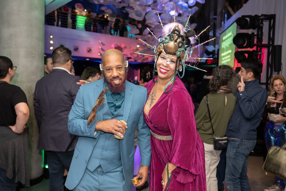 couple posing together at a party smiling. L - man in blue suit, R - woman in burgandy velvet dress with elaborate, tall headdress