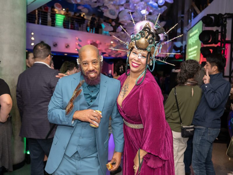 couple posing together at a party smiling. L - man in blue suit, R - woman in burgandy velvet dress with elaborate, tall headdress