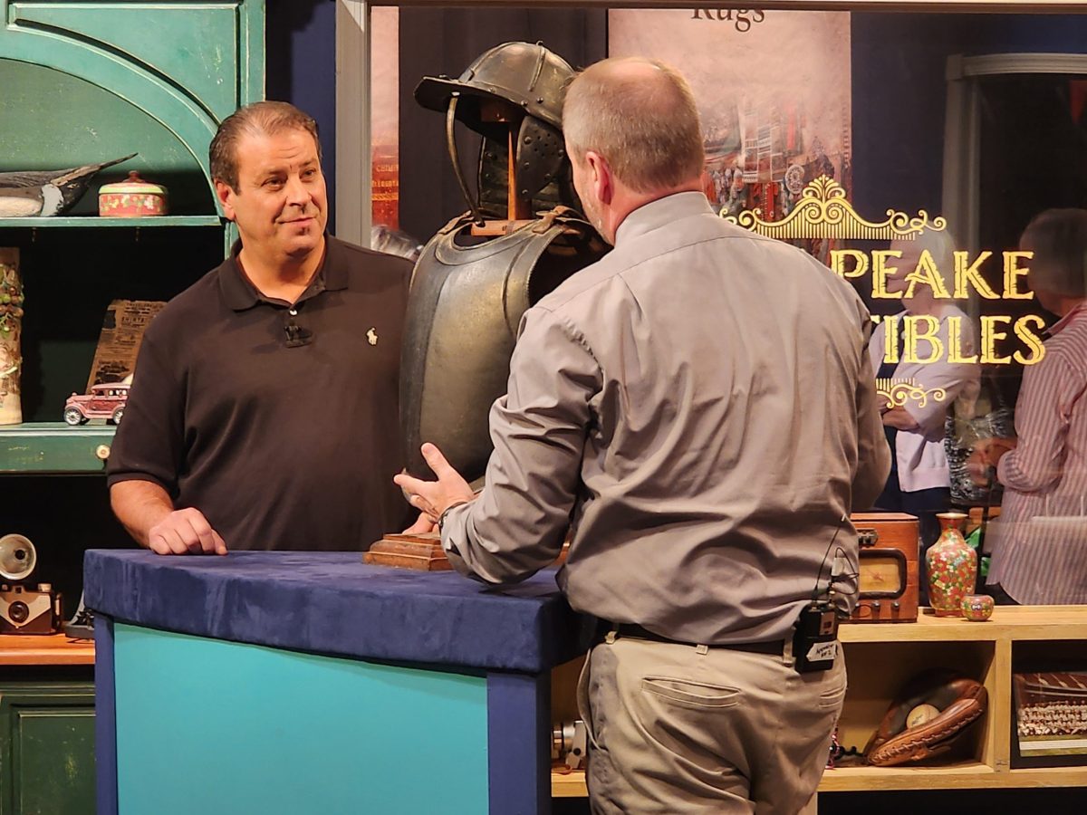 two men at a table on a tv set with a partial set of armor on the table