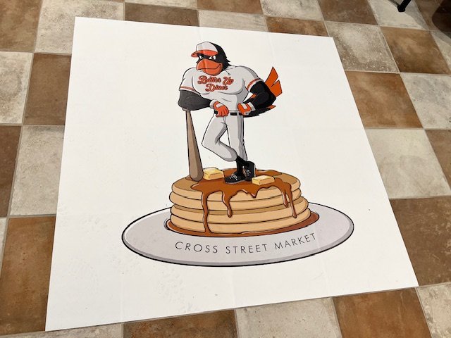 An oriole bird dressed in Baltimore Orioles baseball uniform with "Batter Up Diner" written on the chest. The bird leans on a baseball bat on top of a stack of pancakes. The pancakes sit on a plate with "Cross Street Market" written on it.