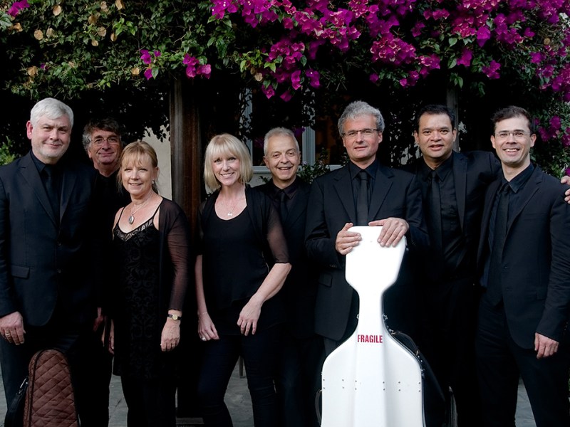 seven musicians stand next to each other dressed in black, one holding a white cello case balanced on the ground