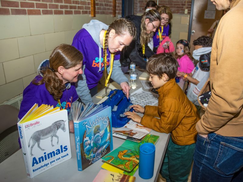 A group of people lean over a table. Two See Beyond Volunteers talk with a child as the child, accompanied by an adult, pushes the keys of a braille writer. The table displays a couple of books that include braille translation and descriptions.