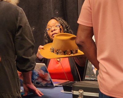 woman sitting at table in red shirt and glasses holding a hat with brim