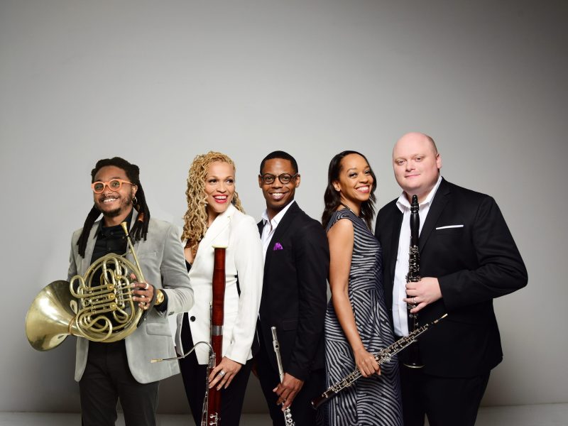 3 women and 2 men standing next to each other smiling and holding wind instruments against a gray backdrop