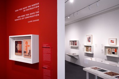 Artworks are displayed in the newly unveiled Irene and Richard Frary Gallery at Johns Hopkins University's Hopkins Bloomberg Center in Washington, D.C. In the foreground, artwork hangs in a glass box against a red wall. Text on that wall reads "Here you have the heroes of destruction / and here you have the fanatics of construction. Lajos Kassák." Additional art hangs on a white wall in the background.