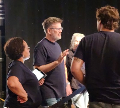 man in black shirt and glasses talking with 3 people, black background behind them