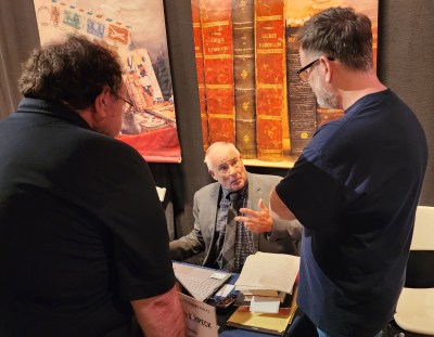 two men standing facing a man sitting down, discussing manuscripts on a table between them.