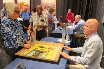 (l) woman standing talking to man (r) seated about painting between them on table