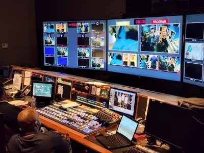 TV control room with multiple screens and control board, and two people sitting behind board.