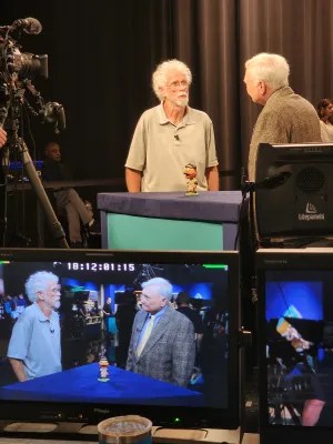 two men speaking with podium between them on which is an old Orioles Bird bobblehead, with screen filming them on bottom half of photo