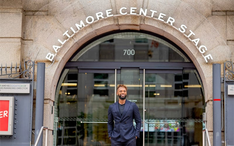 Man stands in black suit smiling under arched doorway with words Baltimore Center Stage in letters above arch in white