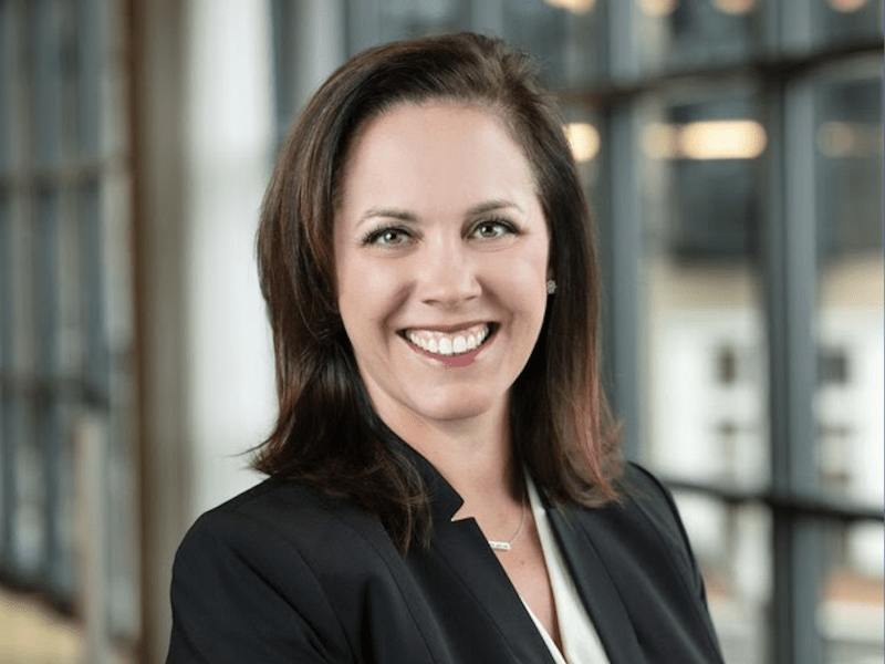 Woman with dark hair looking at camera smiling, wearing black suit jacket over white shirt.