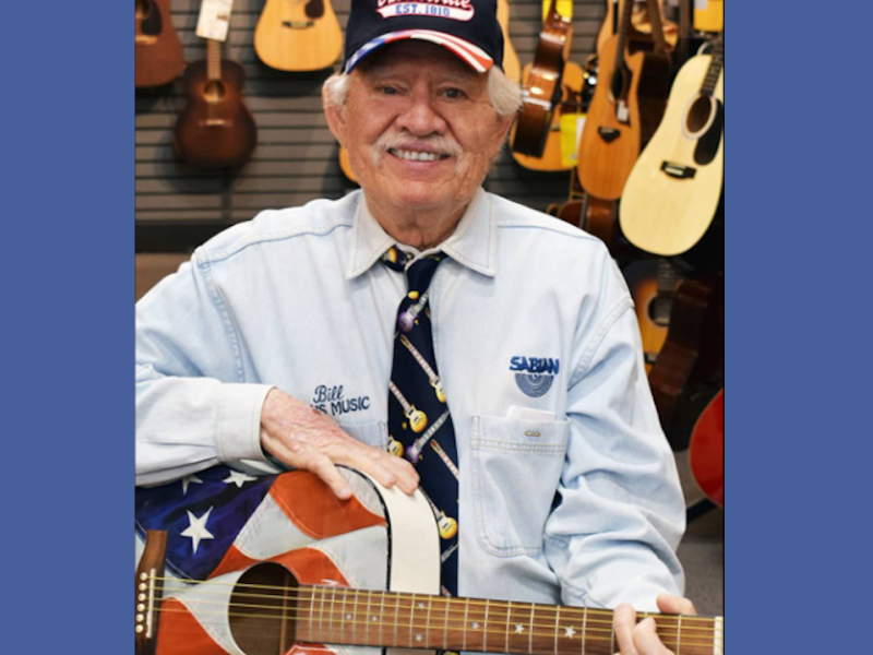 Man in blue shirt and tie wearing a baseball cap holding an American flag themed guitar, smiling at camera