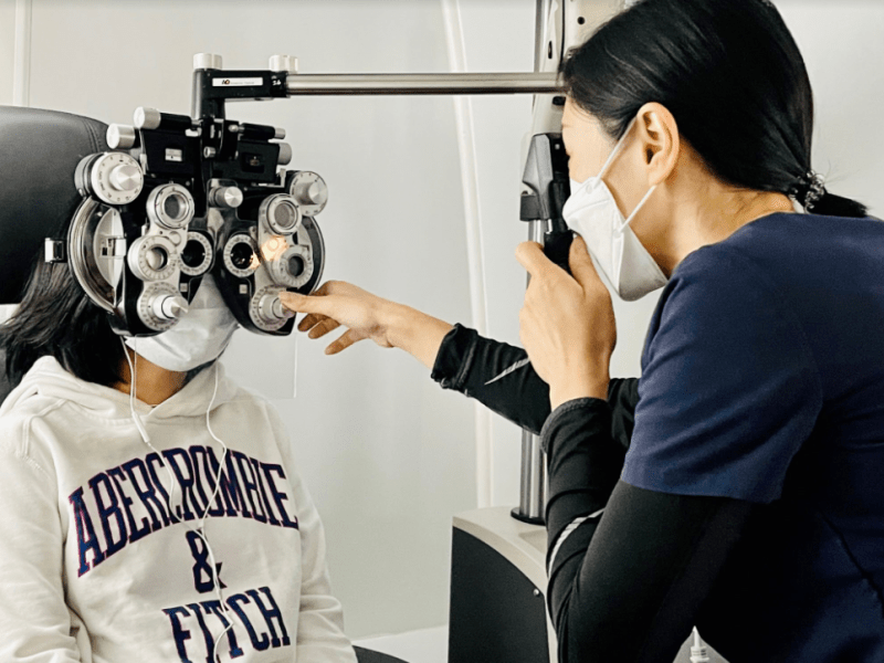 adult woman in dark shirt and white mask gives eye exam to older child wearing a gray Abercrombie and Fitch sweatshirt and a mask.