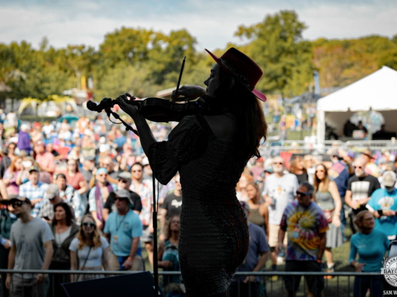 Silhouette of woman in cowboy hat playing violin on stage with crowd standing beyond in audience in an outdoor setting