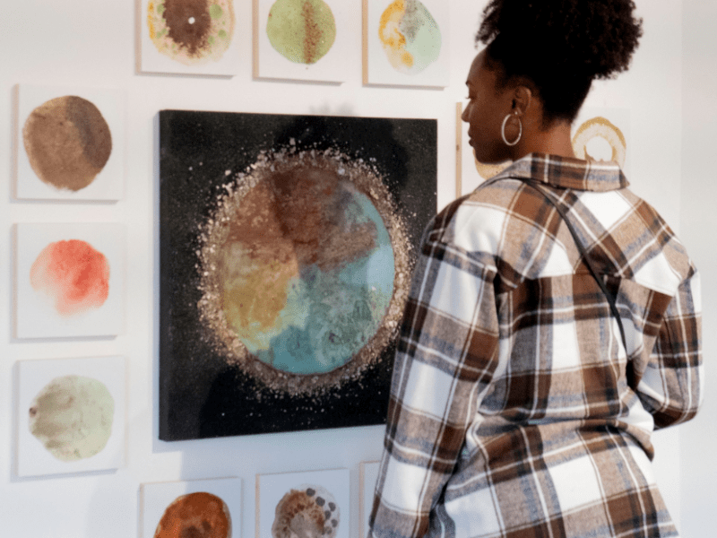 Woman in plaid shirt looking at cluster of paintings on a white wall