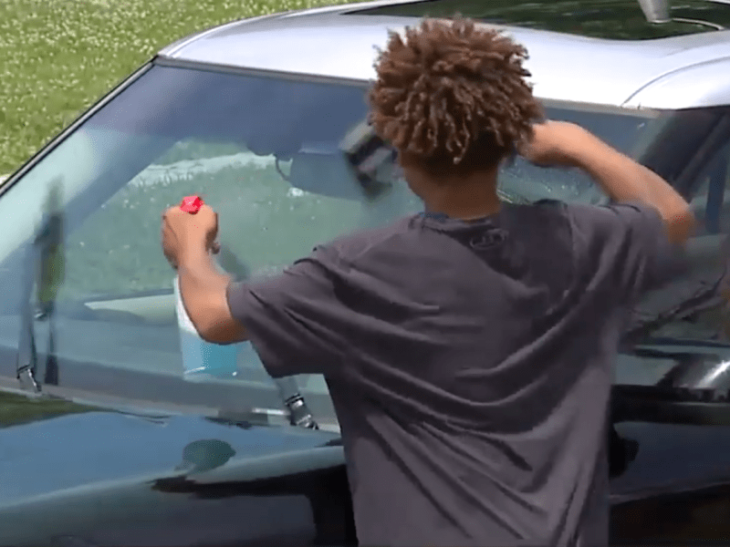 Screenshot of young boy from behind cleaning the windshield of a car