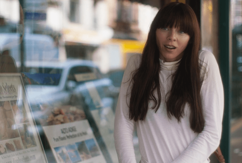 woman with long brown straight hair and bangs, wearing a long sleeved white sweater shrugging her shoulders outside a newsstand/store