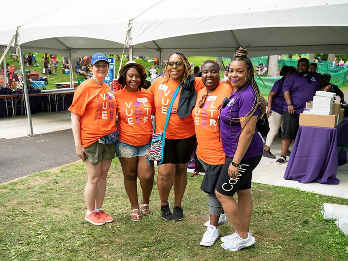 Five women arm in arm, four wearing orange "volunteer" shirts, the last wearing a purple shirt, standing outside in front of a white pop up tent