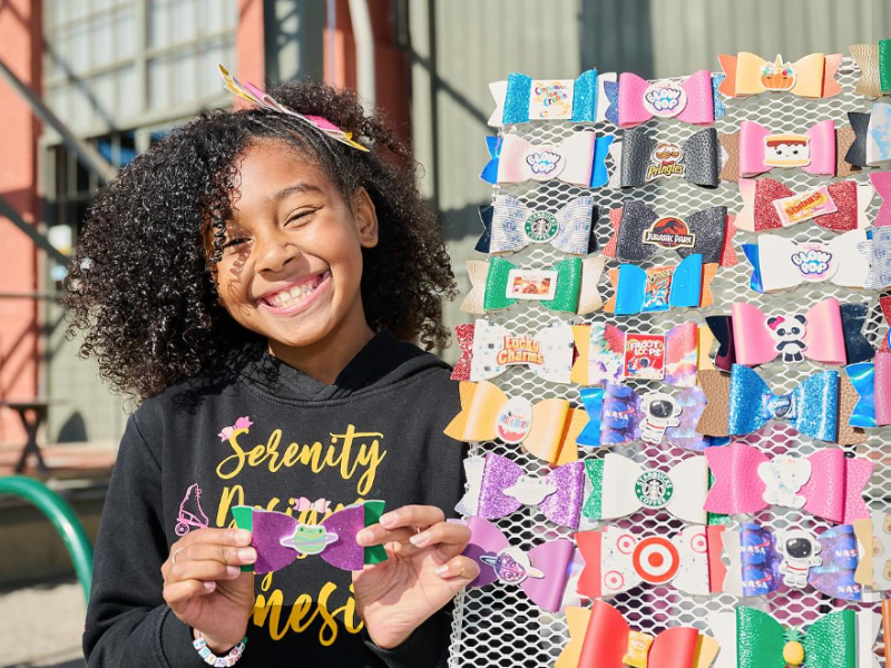 girl with black curly hair smiling and holding a handmade bow, next to a small rack of more handmade bows for hair