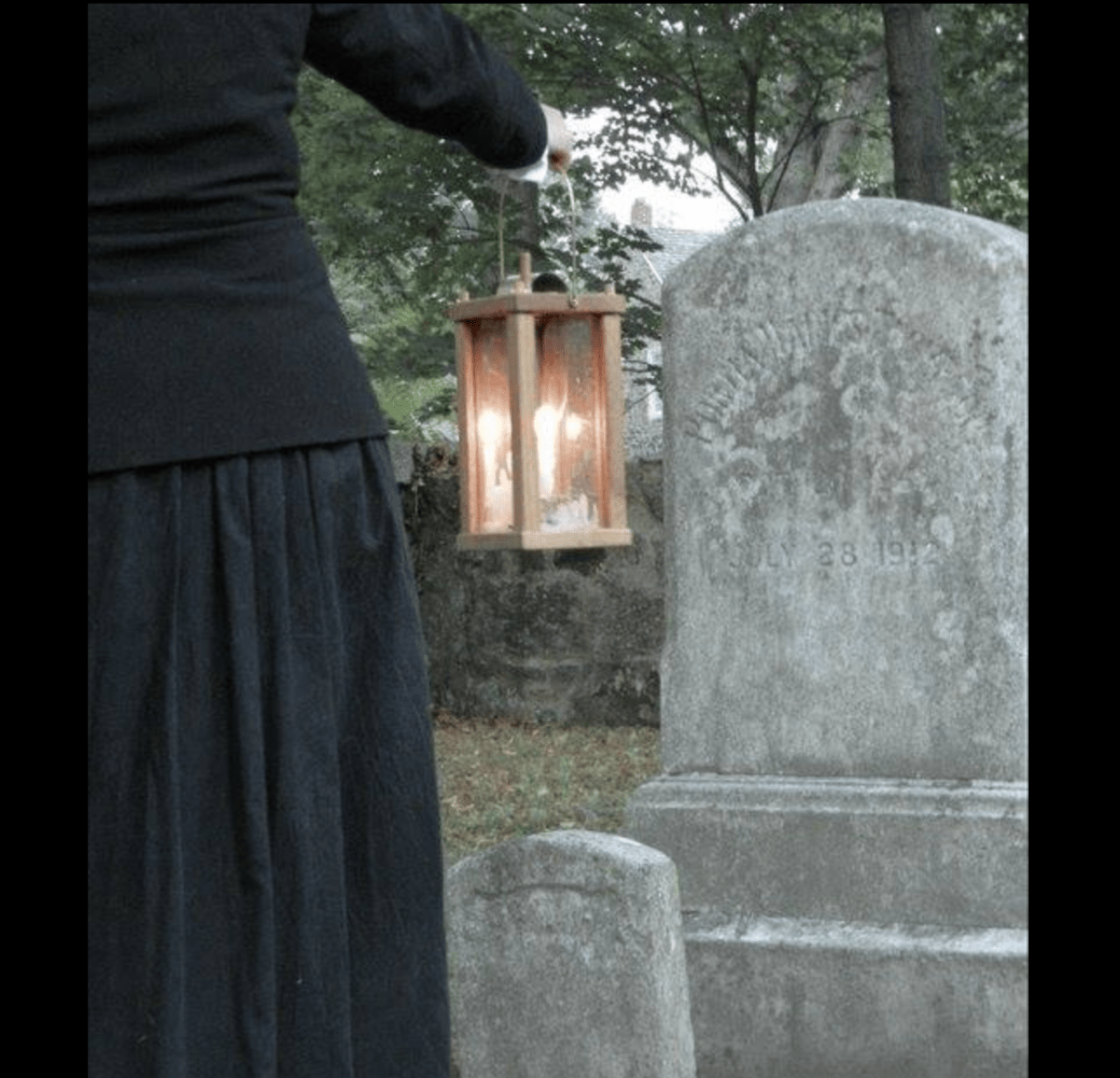 partial shot of woman from behind in long black skirt holding lantern looking at a gravestone