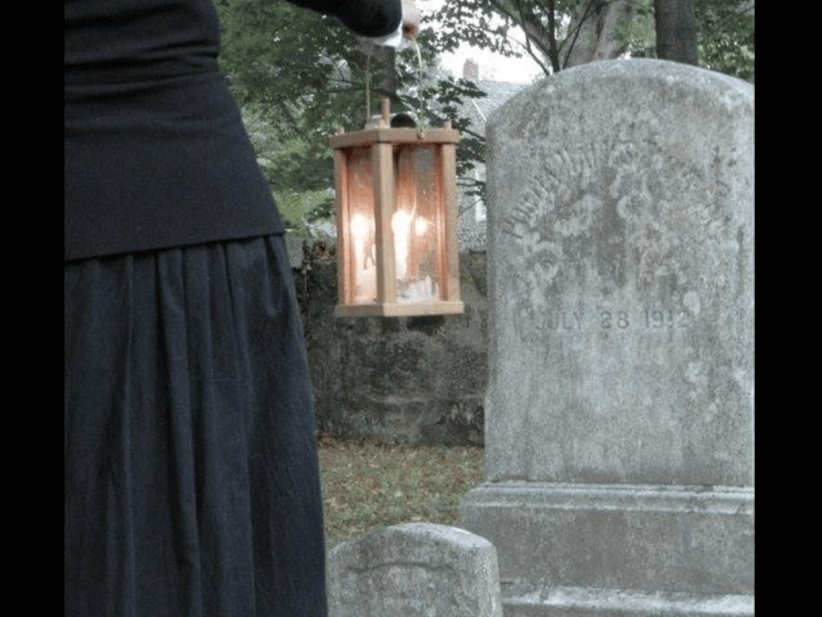 partial shot of woman from behind in long black skirt holding lantern looking at a gravestone