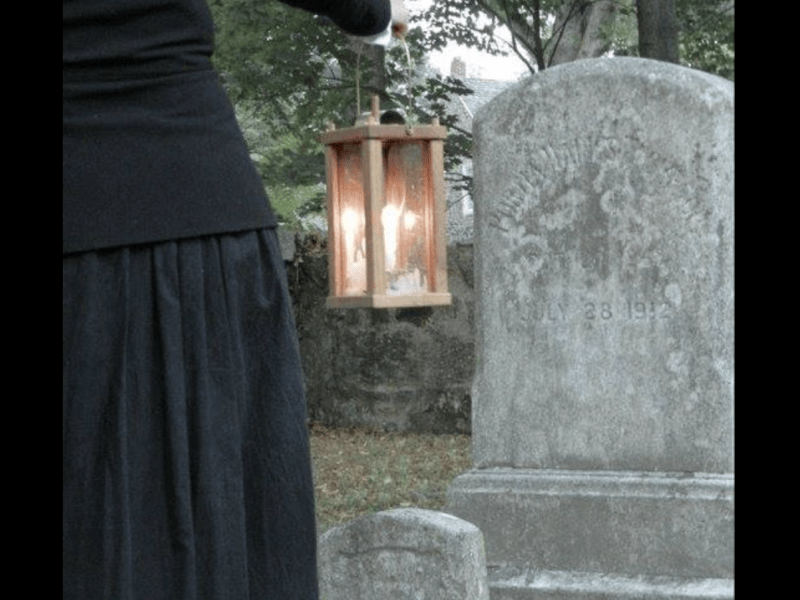 partial shot of woman from behind in long black skirt holding lantern looking at a gravestone