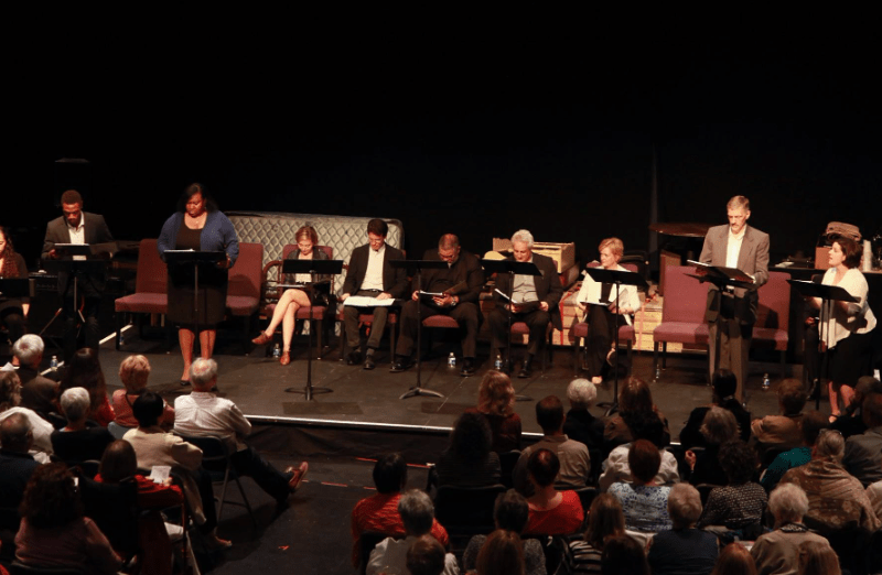 Actors on a stage doing a read through of a script sitting on folding chairs with audience looking on