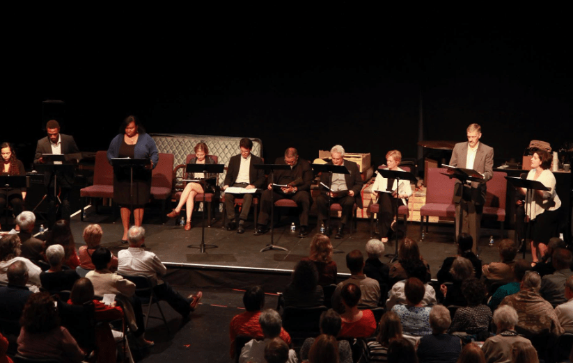 Actors on a stage doing a read through of a script sitting on folding chairs with audience looking on