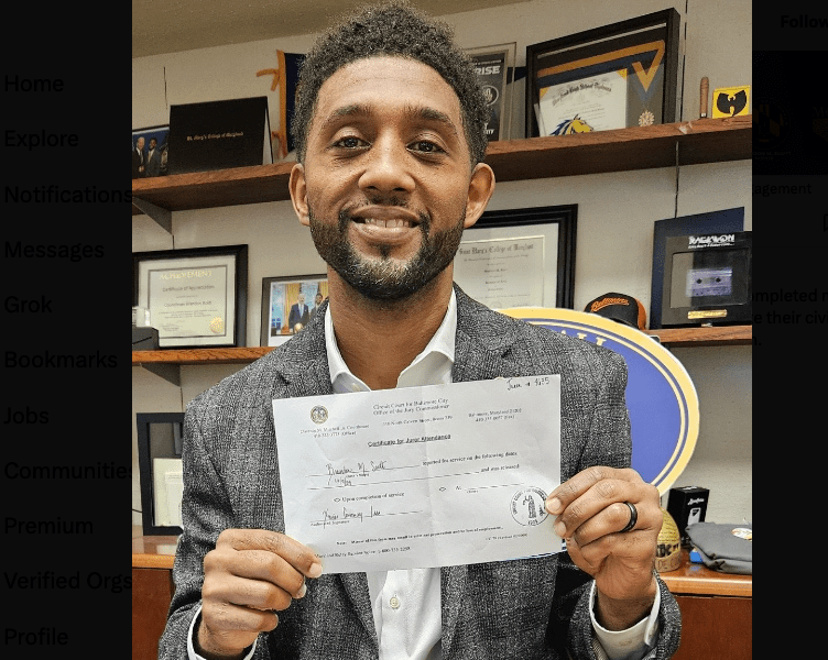 Mayor Brandon Scott seated at desk holding form and smiling at camera