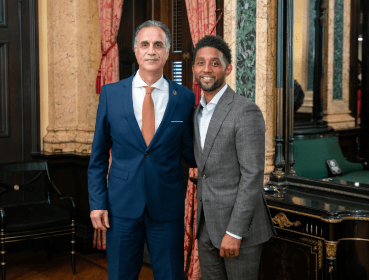 two men in suits and ties stand side by side smiling in front of a curtain