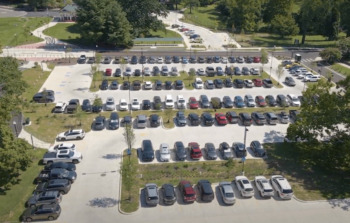 skyshot of parking lot with white pavement and rows of cars with grass and trees surrounding it