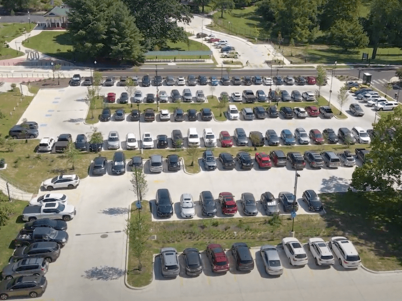 skyshot of parking lot with white pavement and rows of cars with grass and trees surrounding it