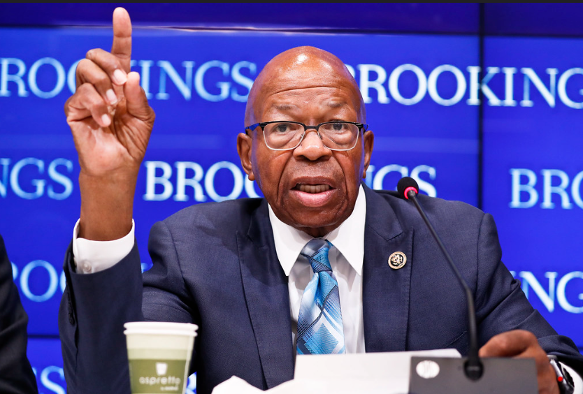 Man sitting at table wearing suit, tie, glasses, holding his hand up with one finger pointed. Blue banner behind him says "BROOKINGS" repeatedly.