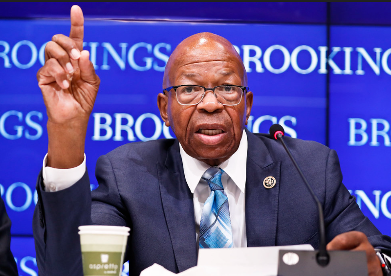 Man sitting at table wearing suit, tie, glasses, holding his hand up with one finger pointed. Blue banner behind him says "BROOKINGS" repeatedly.