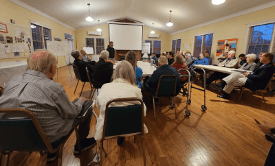 man stands at table where people sit in community room and others sit in chairs around the room